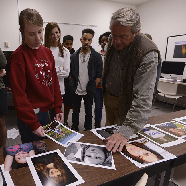 Professor and student critiquing photographs scattered on a table, while others look on.