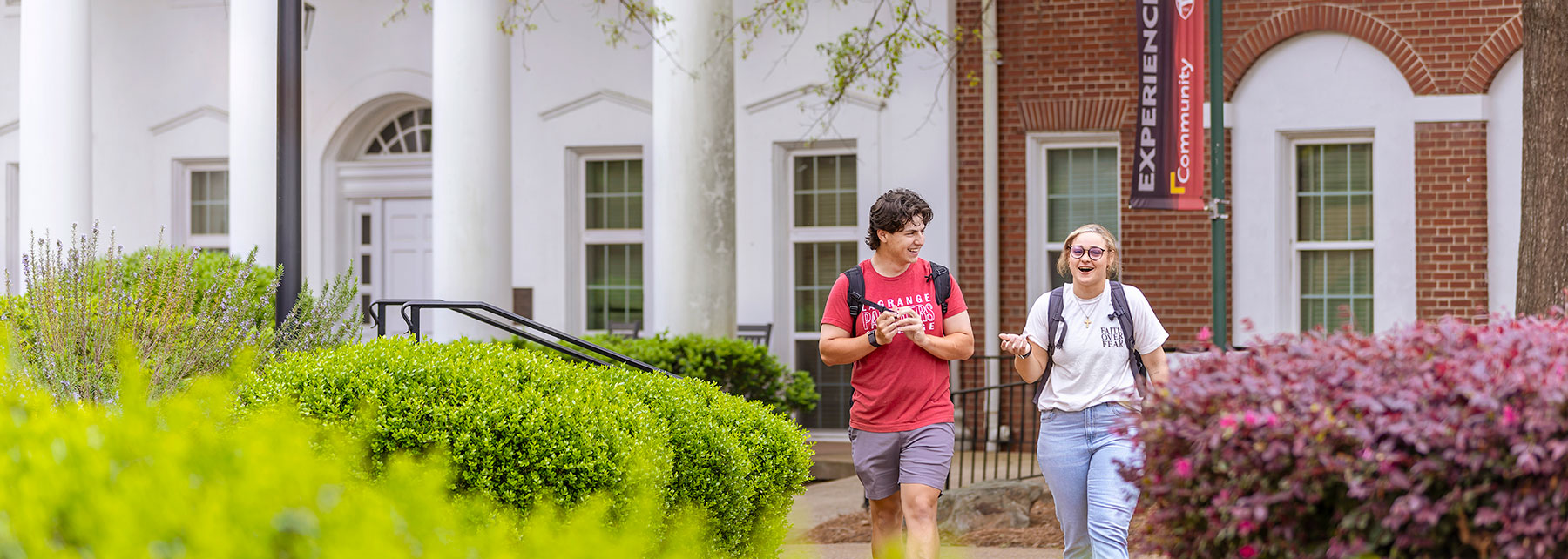 Two students walk along a cobblestone path with a college banner in the background