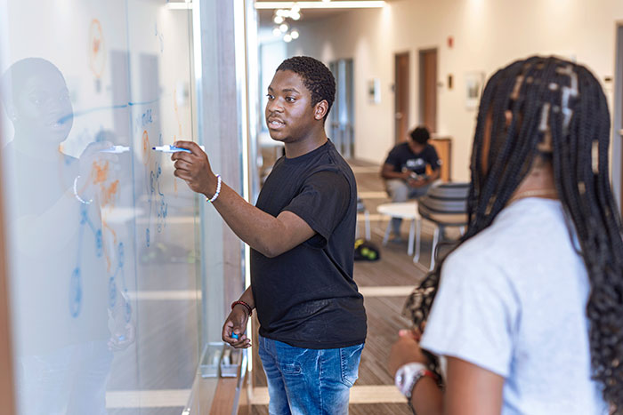 a male student works at a whiteboard while two students stand in the back and foreground