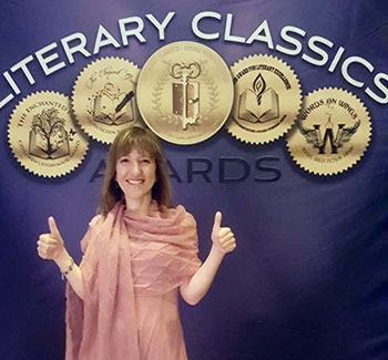 Dr. Patricia Marchesi gives two thumbs up while standing in front of a Literary Classics Awards banner