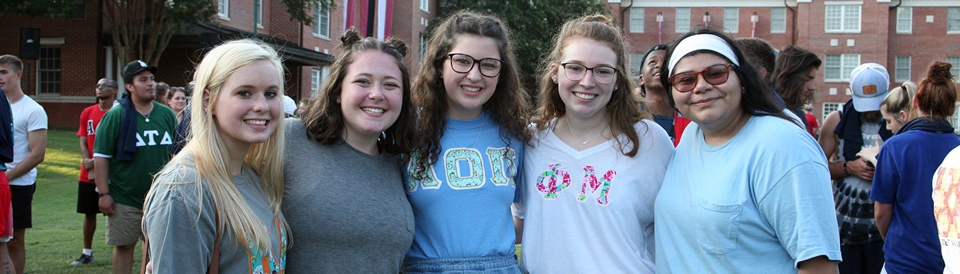 Five students shoulder to shoulder facing the camera in a crowd of other students