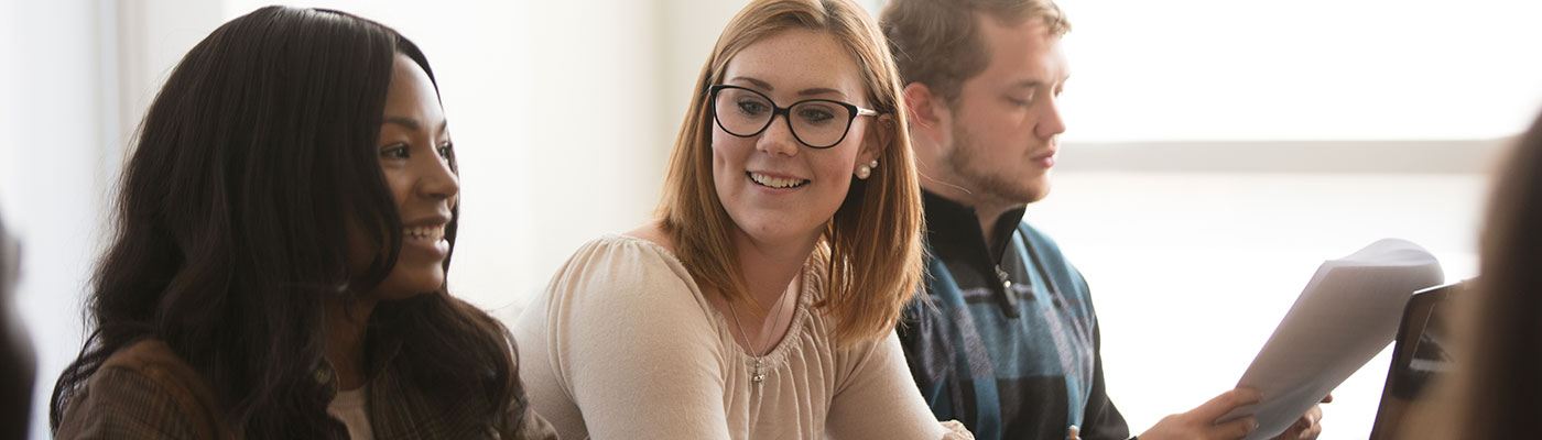 Three students at a table facing the forward. One is reading notes, the other two are chatting.