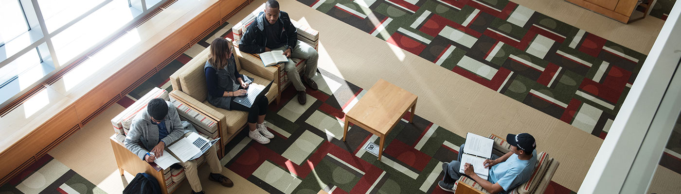 Overhead view of four students on comfy chars studying in the library lobby. Two of them are actively interacting.