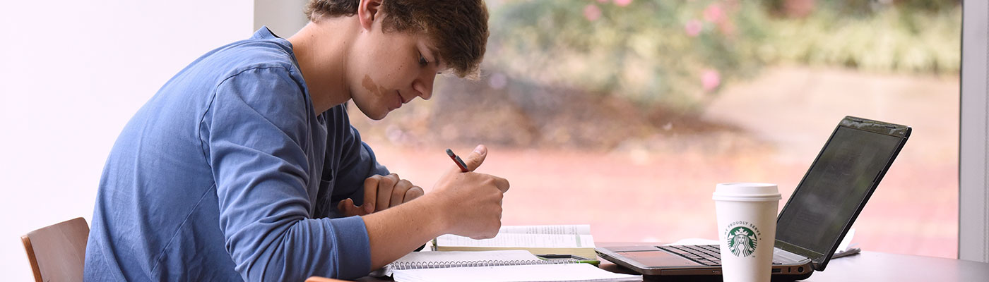 A young male student sits at a high top table working on a laptop in a sunlit room