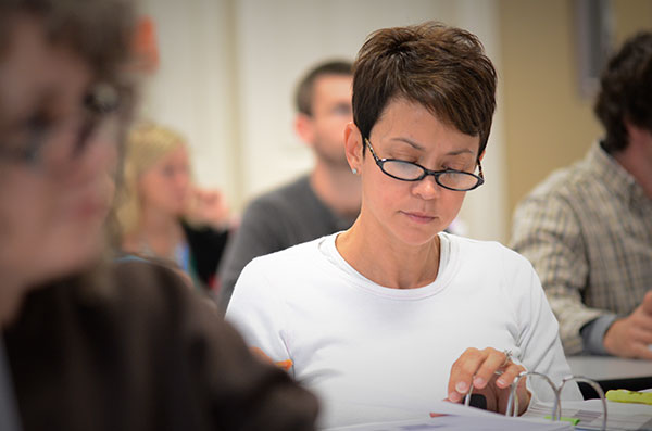 Female student in a white shirt works on a paper at a desk in the middle of a classroom