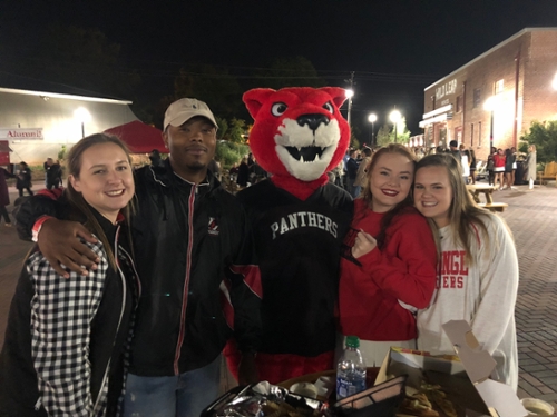 {Four people stand with Pouncer at an evening alumni event}