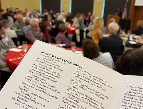 {A scholarship luncheon program in the foreground with a group of guests in the background}