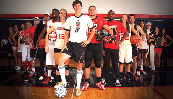 student athletes from multiple sports stand in a V-formation in the gym holding various sporting equipment
